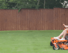 a person is riding an orange lawn mower on a lush green lawn in front of a wooden fence .