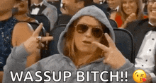 a woman wearing sunglasses and a hoodie is sitting in a crowd of people and says wassup bitch !