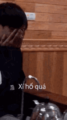a man covering his face with his hands in front of a wooden wall with the words xi ho qua on the bottom