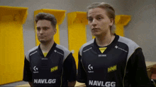 two men wearing navi.gg shirts stand in a locker room