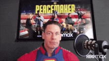 a man in a superman costume is standing in front of a microphone in front of a poster for peacemaker .