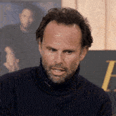 a man with a beard wears a black turtleneck sweater with the letter h in the background