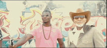 a man wearing sunglasses and a fur hood stands in front of a wall with graffiti