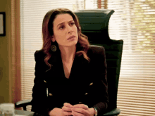 a woman in a black jacket sits in a chair in front of a window with blinds