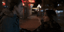 two women are standing on a sidewalk at night with a red sign that says ' a ' on it