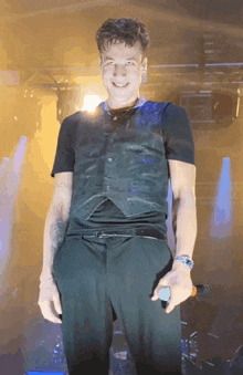 a man in a black vest and black pants is holding a microphone and smiling