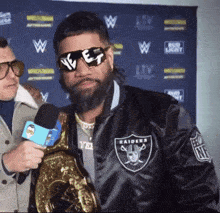 a man wearing sunglasses and a raiders jacket is holding a microphone