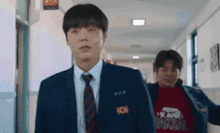 a man in a suit and tie is walking down a hallway with another man wearing a baksal shirt .
