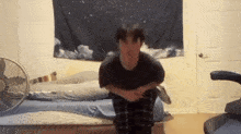 a man is squatting on a bed in a bedroom with a fan and a tapestry hanging on the wall .