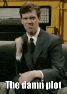 a man in a suit and tie is sitting on a couch with the caption `` the damn plot '' .
