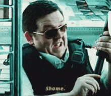 a man wearing sunglasses is holding a gun and saying shame .