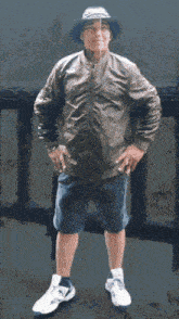 a man wearing a hat and a brown jacket stands with his hands on his hips