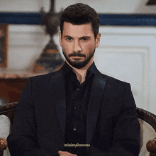 a man with a beard wearing a black suit and black shirt is sitting in a chair