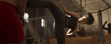a woman in a black top is kicking a man in red shorts in front of a chain link fence