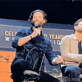 a man speaking into a microphone in front of a wall that says celeb