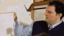 a man in a plaid shirt and tie is pointing to a white board .