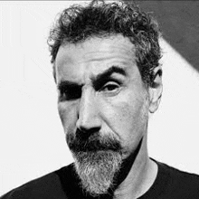 a man with a beard and curly hair is wearing a black shirt and looking at the camera .
