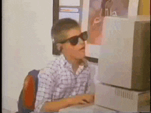 a boy wearing sunglasses sits in front of a computer monitor