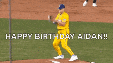 a baseball player in a yellow uniform is standing on the pitcher 's mound and saying `` happy birthday aidan ! ''