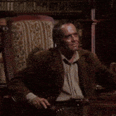 a man in a brown jacket is sitting in a chair in front of a bookshelf
