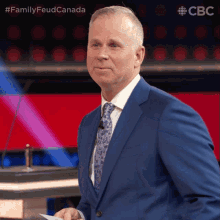 a man in a blue suit and tie is standing in front of a microphone with the words family feud canada behind him