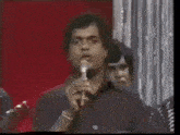 a man is singing into a microphone while another man looks on .