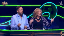 a man and a woman are sitting at a table with the masked singer on the screen