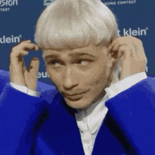 a man with blonde hair is wearing a blue suit and adjusting his earring .