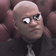 a bald man wearing sunglasses and a suit looks at the camera