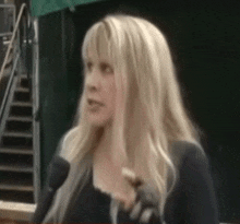 a woman with long blonde hair is talking into a microphone while wearing black gloves