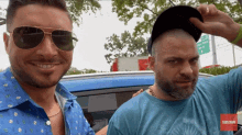 a man wearing sunglasses and a hat stands next to another man wearing a blue shirt