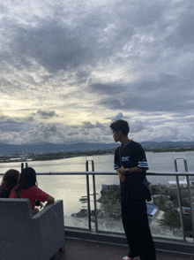 a man wearing a shirt that says tbc stands on a balcony overlooking a river