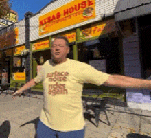 a man in front of a kebab house wearing a yellow shirt that says surface noise rides again