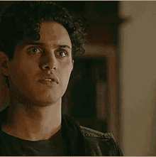 a young man with curly hair is wearing a black shirt and a leather jacket and looking at the camera .