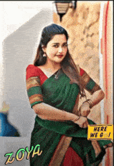 a woman in a green and red saree stands next to a sign that says here we go
