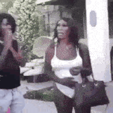 a woman in a white tank top is holding a black purse while standing next to a man .