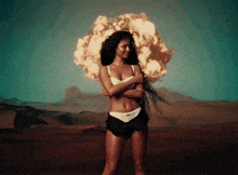 a woman in a white bra and black shorts stands in front of an explosion in the desert