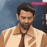 a man with a beard is wearing a brown jacket with a pinkvilla logo in the background