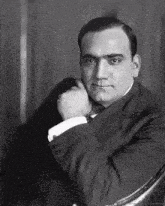 a black and white photo of a man in a suit and tie sitting in a chair with his hand on his chest .
