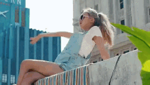 a girl wearing overalls and sunglasses is sitting on a ledge with her arms outstretched