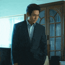 a man in a suit stands in front of a bookcase with his hands in his pockets
