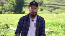 a man with a beard is standing in a field wearing a hat .