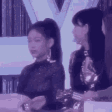 two women are sitting next to each other in a room . one of the women is wearing a ponytail .