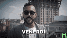 a man with a beard wearing sunglasses is standing in front of a building and saying venerdi .