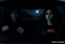 a man is driving a car next to a woman who is wearing a wanted shirt