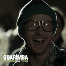 a man wearing glasses and a green hat is smiling in front of a poster for the guarimba international film festival