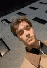 a man in a tan coat is standing in front of a building and making a funny face .