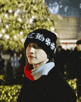 a young man wearing a black hat and a red scarf is standing in front of a christmas tree .