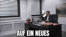 a man in a suit is sitting at a desk in an office with the words auf ein neues written above him .
