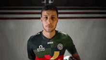 a man wearing a jako jersey holds a soccer ball